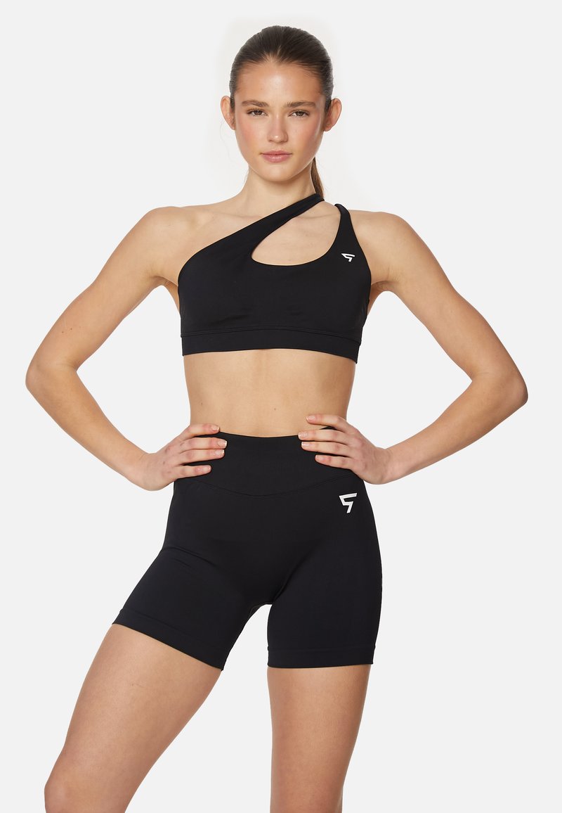 Črn športni nedrček ima asimetričen dizajn s izrezom in usklajenimi visokimi kratkimi hlačami. Narejen je iz raztegljivega, gladkega materiala.