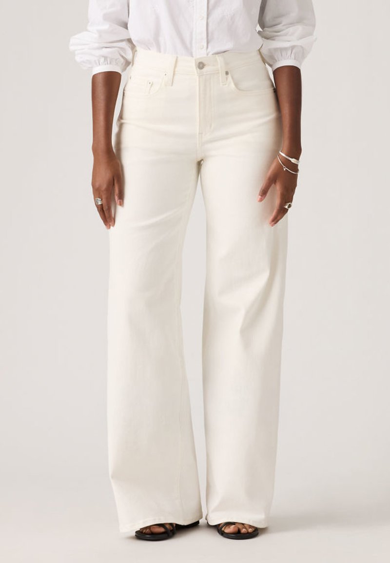 Γυναίκα που φοράει λευκά τζιν παντελόνια με ψηλή μέση και ευρύ πόδι, λευκό πουκάμισο με κουμπιά, μαύρα σανδάλια και ασημένια δαχτυλίδια και βραχιόλια.