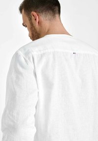 Man die met zijn rug naar de kijker staat en een wit, lange mouwen, linnen shirt draagt met een klein gestikt detail op de bovenrug.