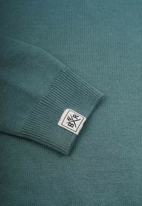 Tropfeneisblau gestrickter Pullover mit gerippten Bündchen; verfügt über eine subtile Textur und ein weißes Etikett mit schwarzer Schrift am Bündchen.