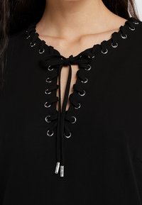Czarna bluzka z głębokim dekoltem w kształcie litery V, ozdobiona srebrnymi oczkami i sznurowanym wzorem. Gładki materiał z delikatnym połyskiem i regulowanymi zapięciami.
