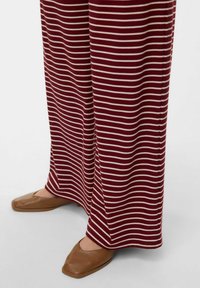 Pantalons rayés bordeaux en texture côtelée, associés à des chaussures plates marron à bout carré. Les rayures horizontales sont prédominantes.