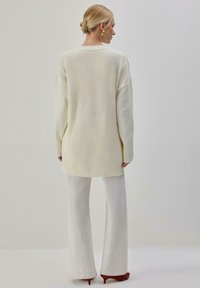 Pull long et oversize crème avec texture côtelée et épaules tombantes, associé à un pantalon blanc à jambes larges et des chaussures à talons rouges.
