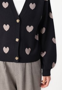 Cardigan nero con motivi a cuore taupe, design a scollatura a V e tre bottoni dorati. Materiale in maglia testurizzata con polsini a coste.