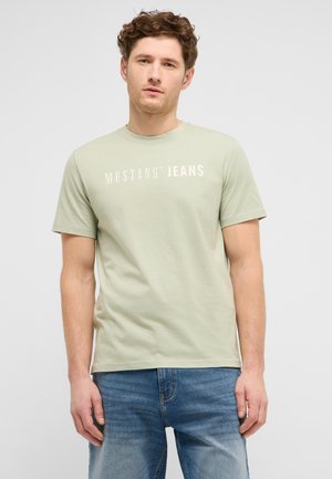 Junger Mann mit lockigem Haar, der ein hellgrünes Mustang Jeans T-Shirt und blaue Denim-Jeans trägt, steht vor einem einfarbigen Hintergrund.