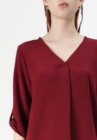 Blusa bordeaux con scollatura a V, tessuto strutturato e maniche arrotolate. Dettaglio di pieghe notevole sul davanti per un design aggiuntivo.