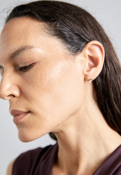 Gul stud ørering med en lille klar ædelsten, båret af en person med mørkt hår. Glat hudstruktur og diskret makeup, set fra en vinkelprofil.
