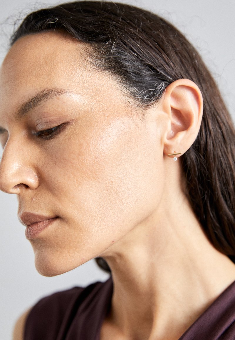 Zlatý náušnice s malým průhledným drahokamem, nošená osobou s tmavými vlasy. Hladká textura pleti a decentní make-up, pohled z úhlu.