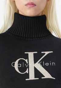 Zwarte ribbel turtleneck trui met een groot beige CK-logo en de tekst "Calvin Klein", gebreide textuur en aansluitend ontwerp.