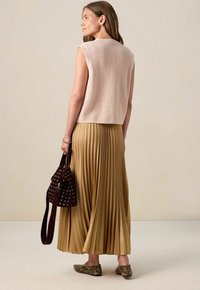 Haut sans manches en ribbed rose clair, jupe plissée dorée, ballerines marron à motifs et un sac seau bordeaux avec des œillets, vu de dos.