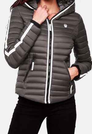 Femme portant une doudoune matelassée gris foncé avec une fermeture éclair blanche et des rayures sur les manches, les mains ajustant le col et dans la poche.