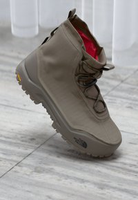 Beige høyhælte tursko med Vibram såle og rød indre fôr, "The North Face"-logo på sålesiden, på en bakgrunn av glatt stein.