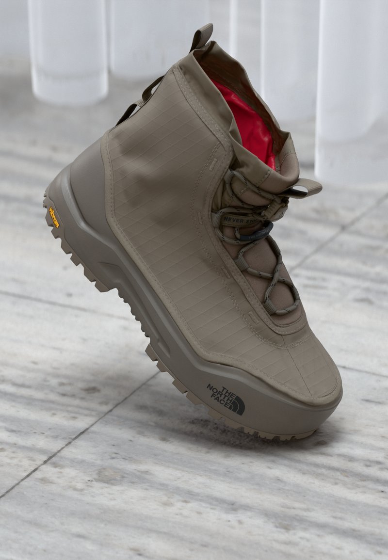 Beige høyhælte tursko med Vibram såle og rød indre fôr, "The North Face"-logo på sålesiden, på en bakgrunn av glatt stein.