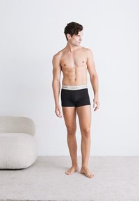 Schwarze Boxershorts mit einem grauen Bund, getragen von einem männlichen Modell mit schlanker Statur, das barfuß auf einem hellen Teppich steht.