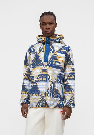Junger Mann mit geflochtenem Haar, der einen mehrfarbigen Kapuzenfleece mit geometrischem Muster und weiße Hosen trägt, schaut mit neutralem Gesichtsausdruck nach vorne.