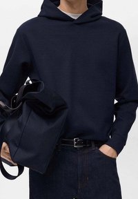 Granatowa bawełniana bluza z kapturem z kieszenią kangurką, zestawiona z czarną torbą tote z miękkiego materiału. Ciemne dżinsy dopełniają stylizację.