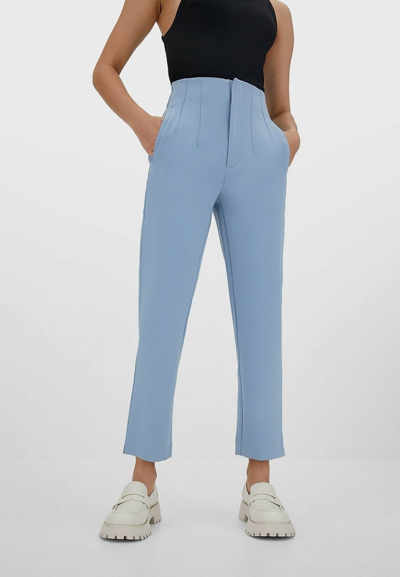 Personne portant un pantalon bleu clair taille haute, un haut noir sans manches, des mocassins blancs épais, debout les mains dans les poches devant un fond uni.