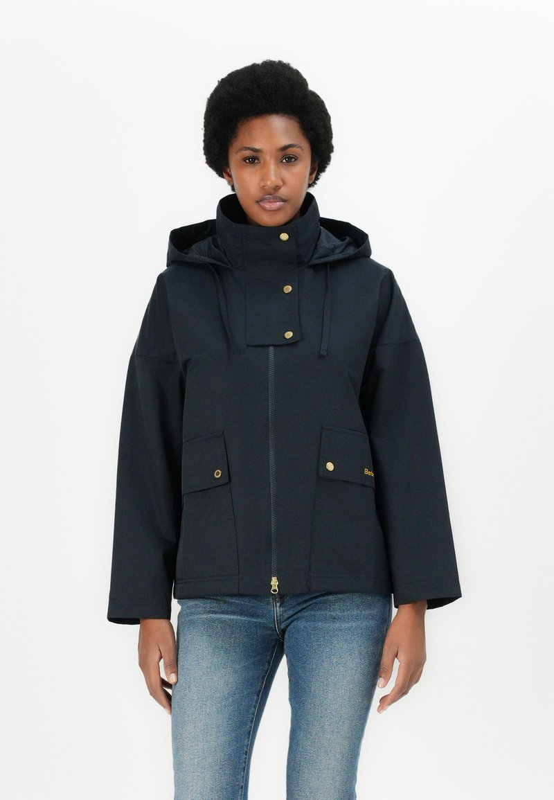 Veste imperméable bleu marine avec un col haut, fermeture éclair frontale et deux grandes poches. Dotée d'accents dorés et d'une capuche. Fabriquée en tissu lisse.