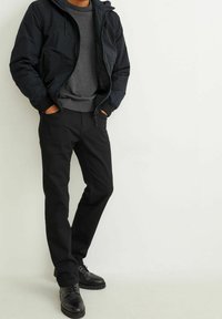 Zwarte zip-up jas met capuchon, grijze sweater en zwarte broek, gecombineerd met zwarte veterschoenen. Over het algemeen een donker kleurenpalet.