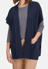 Marinblå stickad cardigan med öppen framsida, två fickor och en avslappnad passform. Under finns en mönstrad blus med rund hals och trekvartsärmar.