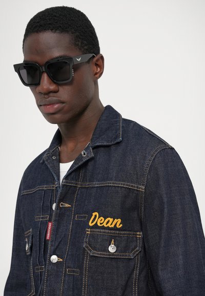 Mørkeblå denimjakke med gult broderet navn "Dean", sølvknapper og klassisk krave. Sorte solbriller med vinklet design.