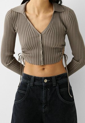 Top crop côtelé marron avec décolleté en V et fermeture à boutons ; il présente des manches longues, des liens sur les côtés et une coupe ajustée. Associé à un jean noir.