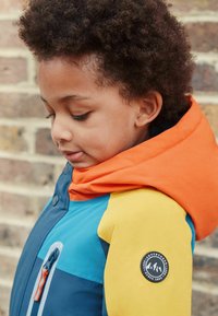 Kleurrijke kinderjas met een oranje capuchon, een blauw en gele romp, uitgerust met een rits, drukknoopjes en een ronde logo op de mouw.