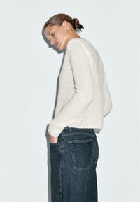 Maglione lavorato a maglia beige chiaro con una finitura testurizzata, abbinato a jeans a vita alta blu scuro. Il maglione è accorciato in vita.