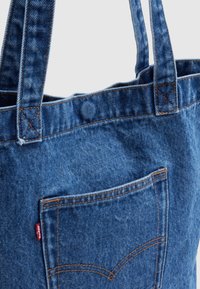 Τσάντα ταχύτητας από μπλε τζιν με εμφανή μπροστινή τσέπη, διπλούς ιμάντες ώμου και πορτοκαλί ραφές σε ανοιχτό φόντο.