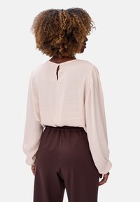 Lichtroze blouse met lange mouwen, voorzien van een sleutelgatdetail aan de achterkant. Gepaard met bruine broeken met elastische taille voor een ontspannen pasvorm.