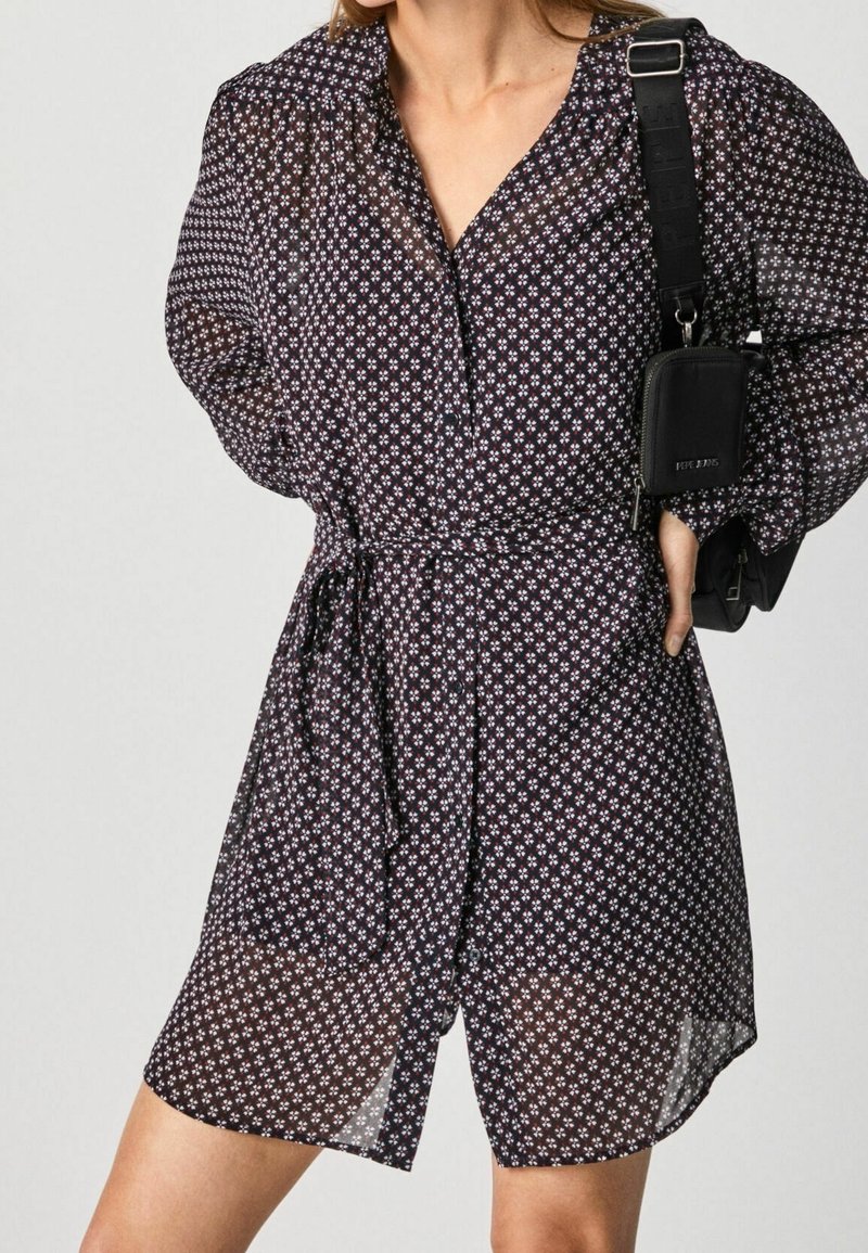 Femme portant une robe-chemise noire à motifs longueur genoux avec une ceinture à la taille et un sac bandoulière noir sur l'épaule.