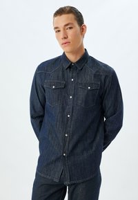 Jeansskjorta i mörkblått med knapplist, två bröstfickor och kontrasterande sömmar. Har långt ärm och en avslappnad passform.