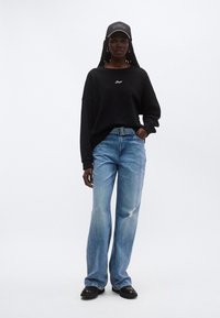 Μαύρο φαρδύ φούτερ με λευκό λογότυπο, ανοιχτό μπλε ξεφτισμένο τζιν παντελόνι και μαύρα παπούτσια, συνοδευόμενα από μαύρο καπέλο με το κείμενο «BOSS».