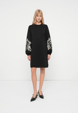Femme debout portant une robe noire à mi-longueur avec une broderie florale blanche sur les manches et des talons aiguilles noirs.