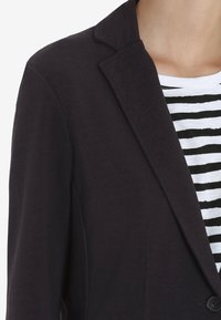 Blazer nero con una finitura testurizzata e rever a scudo, dotato di chiusura a un bottone e indossato sopra una camicia a righe.