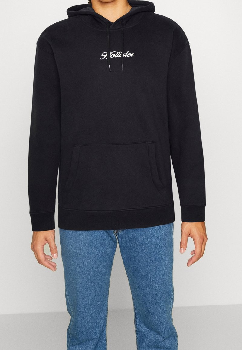 Svart hooded sweatshirt med en framficka och vitt broderat logotyp. Har dragsko och ribbade manschetter, bärs med blå jeans.