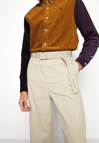 Camisa de pana con secciones marrones y negras, cuello abotonado y botones blancos. Pantalones beige con un cinturón texturizado y detalle del logo.