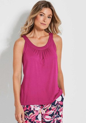 Femme aux longs cheveux blonds portant un haut sans manches magenta et un pantalon à motifs floraux avec des fleurs roses et blanches sur fond marine.