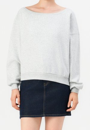 Personne portant un sweat-shirt oversize gris clair avec des manches bouffantes et une mini-jupe en jean bleu foncé sur un fond uni.