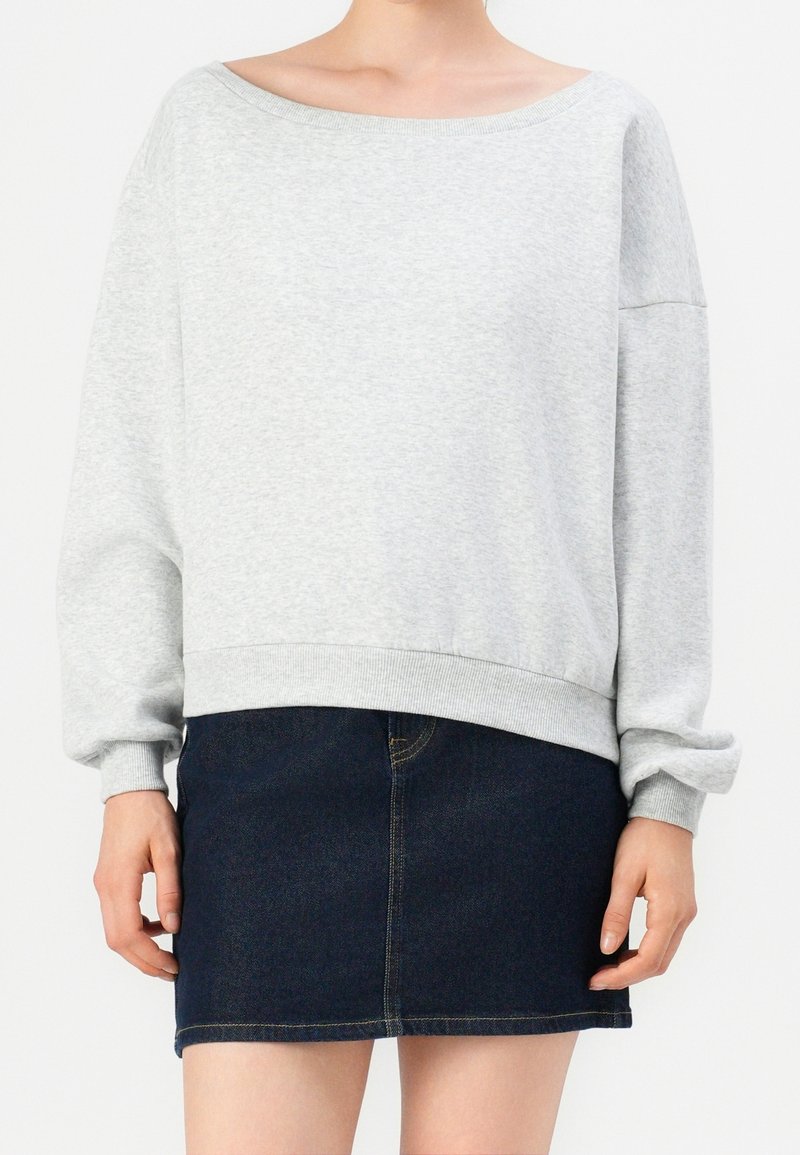 Personne portant un sweat-shirt oversize gris clair avec des manches bouffantes et une mini-jupe en jean bleu foncé sur un fond uni.