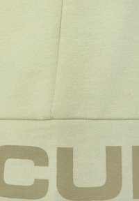 Light green sweatshirt fabric with a ribbed texture; features large beige text at the bottom. Smooth finish with clean seams.