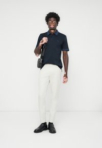 Polo azul-marinho com colarinho em contraste, calças creme e sapatos pretos. O modelo segura uma pequena bolsa preta de ombro, com ferragens prateadas.