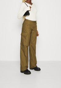 Olivově zelené cargo kalhoty s volným střihem, mnoha bočními kapsami a hladkým povrchem; spárované s přiléhavým bílým dlouhým rukávem a černými botami.