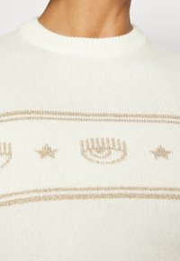 Pull en tricot blanc avec des rayures horizontales dorées, des motifs d'étoiles dorées et un dessin d'œil doré sur la poitrine, sous un col côtelé.