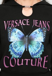 Melna kapuce ar lielu krāsainu tauriņu grafiku, akcentētu ar rhinestones, un tekstu "VERSACE JEANS COUTURE" rozā spīdumā.