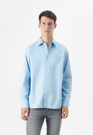 Young man with short hair stands against a plain white background, wearing a light blue button-up shirt and gray jeans, hands relaxed at his sides.