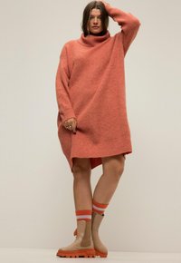 Abito maglione corallo oversize con collo alto, lavorazione a maglia morbida e spacchi laterali. Abbinato a stivali alla caviglia beige decorati con accenti arancioni.