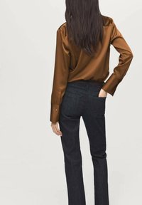 Woman with long dark hair wearing a brown satin blouse and dark straight-leg jeans, standing with one hand in her back pocket.