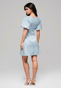 Robe en dentelle bleu clair avec des manches bouffantes, un corsage ajusté et une jupe évasée, mettant en valeur des motifs floraux complexes. Associée à des talons nus à lanières.