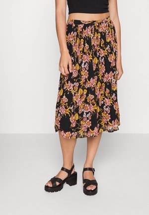 Kvinde iført en sort blomsterprint midi-nederdel med pink og gule blomster, kombineret med sorte chunky hælede sandaler og en sort cropped top.
